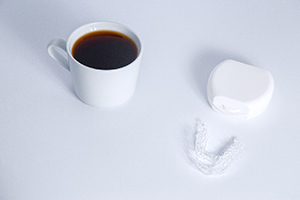 Clear aligners on white surface next to white case and white mug full of coffee